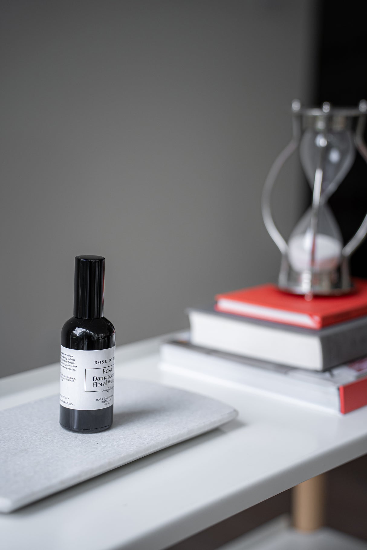 Small black bottle with a label on a gray surface, with books and a hourglass in the background.