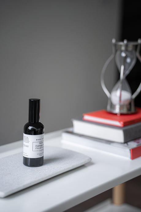 Small black bottle with a label on a gray surface, with books and a hourglass in the background.